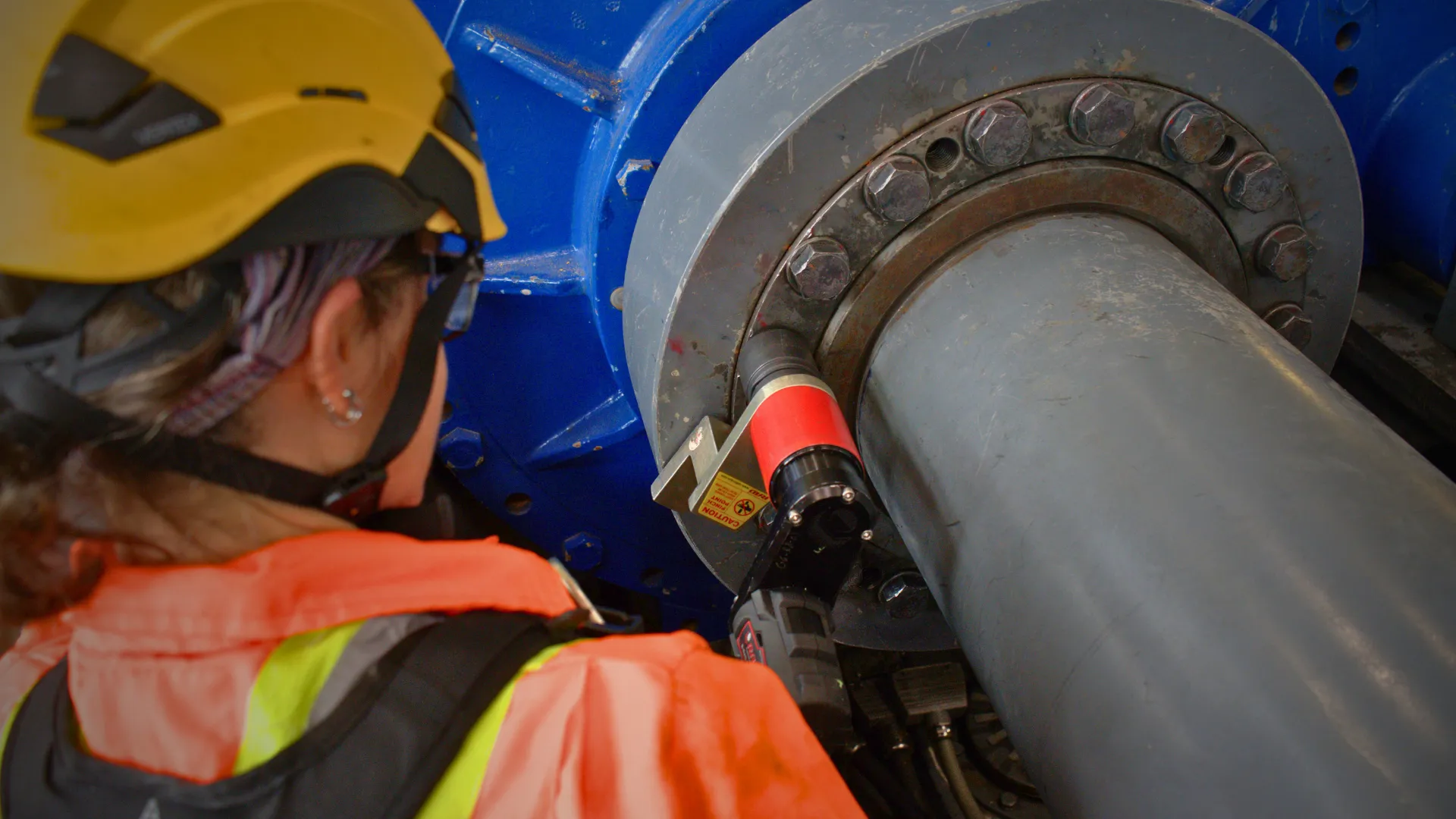 Personne portant casque et lunettes de protection et d'une veste fluorescente observant un très gros boulon d'une éolienne